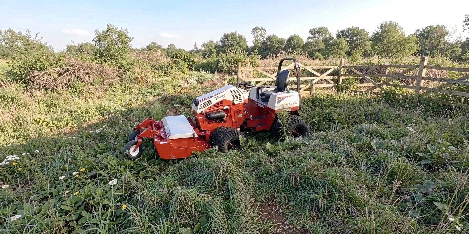 A compact tractor with a rough-cut deck standing in waist-high overgrown vegetation beside a post-and-rail fence.